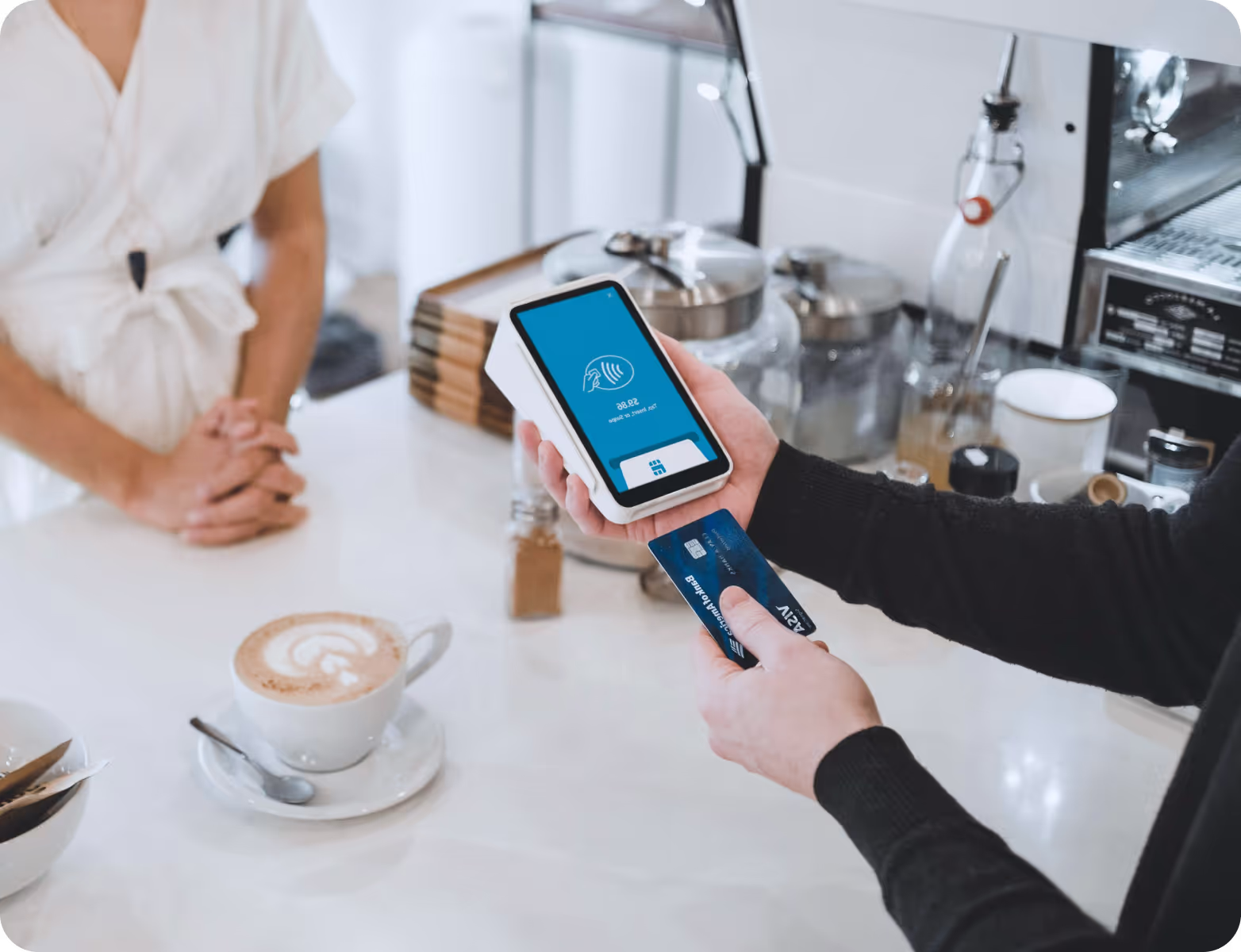 The image shows a person in a café holding a smartphone with a digital payment interface on the screen. The phone is being used to make a contactless payment, as indicated by a card symbol and the text "28.95" on the display. The person is holding a physical card in their other hand, which is partially visible. In the background, a coffee cup with latte art and other items like sugar packets and a coffee machine are visible on the counter. A second person, wearing a white shirt, is standing across from them, likely in the process of completing the transaction.